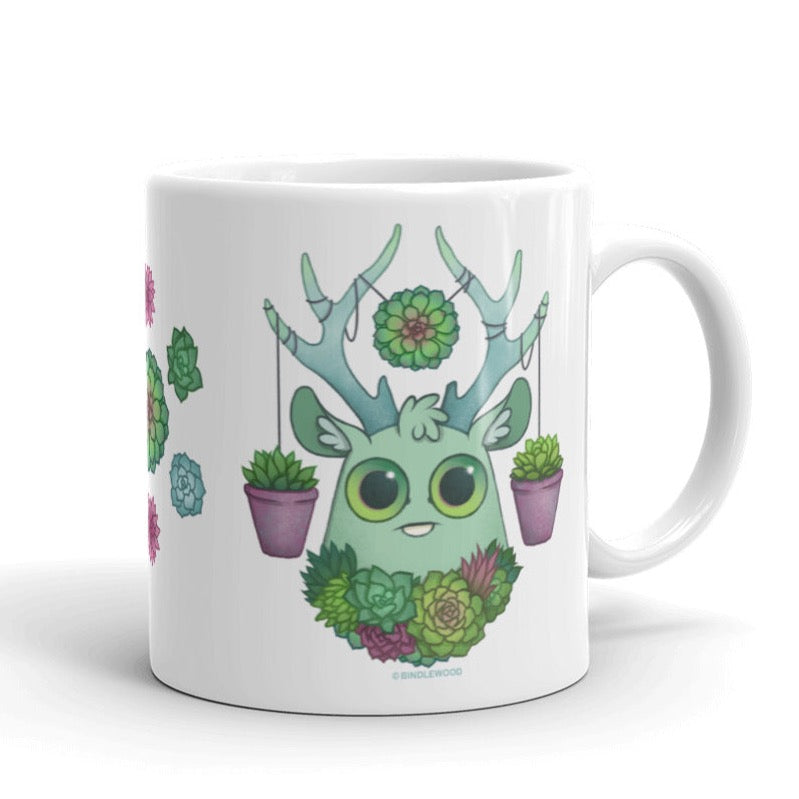 The Succulent Mug by Bindlewood Shop is a whimsical ceramic mug that showcases a cute green creature adorned with large eyes and branch-like antlers. It features an array of succulents and flowers, with pink pots hanging from its antlers, offering a delightful and colorful design that is both charming and dishwasher safe.