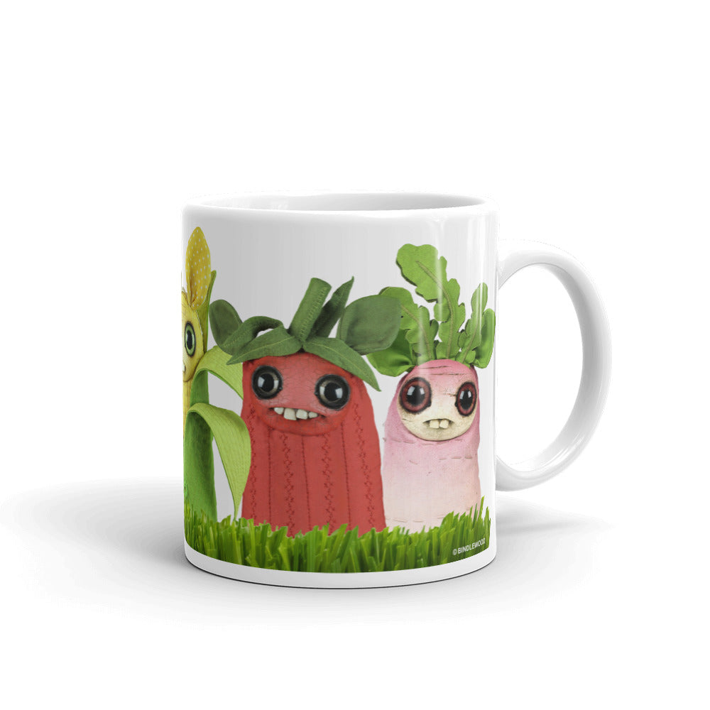 The Bindlewood Shop's Farm Market Dust Bunnies Mug is a white ceramic mug that showcases three adorable, anthropomorphic vegetable characters with smiling faces. The characters depicted are a corn with eyes and leaves, a red radish with large green leaves, and a pink radish with smaller leaves, all set against a grassy background. This charming mug is both microwave safe and dishwasher safe.