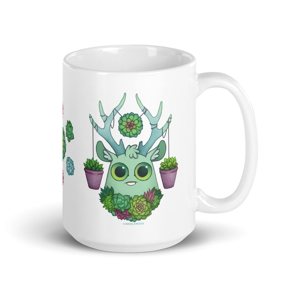 The Succulent Mug from Bindlewood Shop features a charming ceramic design with a cute teal-colored creature adorned with antlers and succulents. The adorable creature boasts large green eyes, and its antlers hold two small potted plants. This whimsical mug is also dishwasher safe for effortless cleaning.